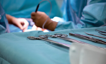 Detailaufnahme von OP-Instrumenten, die auf einem Tisch liegen, im Hintergrund sieht man Hände von Chirurgen, die operieren