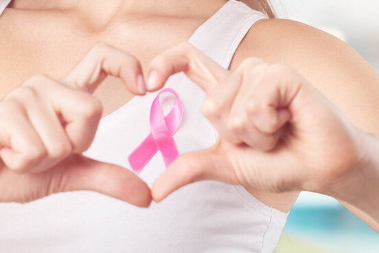 Frau formt mit ihren Fingern ein Herz in Höhe der Brust und rahmt damit eine rosafarbene Schleife ein