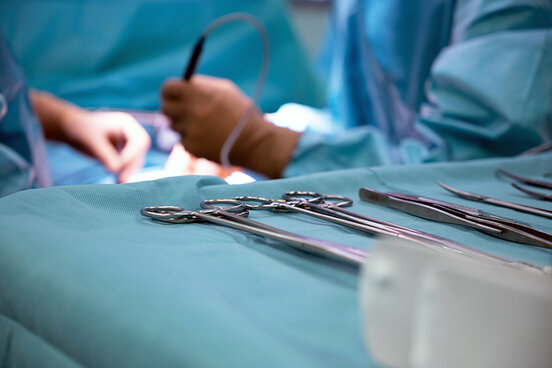 Detailaufnahme von OP-Instrumenten, die auf einem Tisch liegen, im Hintergrund sieht man Hände von Chirurgen, die operieren