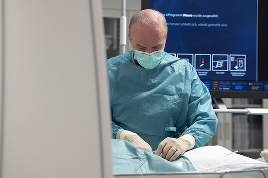 Radiologe, der einen Mundschutz trägt, steht am Tisch einer biplanen Angiographie-Anlage und führt eine Untersuchung mit einem Katheter durch. 