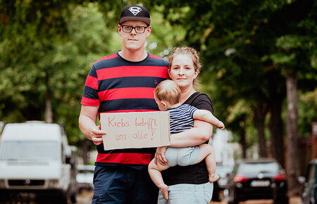 Paar aus Mann und Frau hält ein Kleinkind auf dem Arm. Sie halten ein Schild, auf dem steht "Krebs betrifft uns alle!"